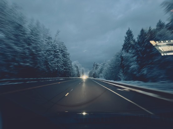 Picture of a bare wet road surrounded by snow covered trees. 
