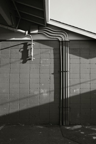 A black and white image showing various metal pipes and ducts attached to a cinderblock wall, with shadows cast on the wall.