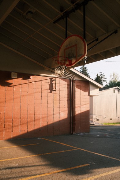 An outdoor basketball hoop attached to a brick building, with pipes and vents running along the underside of the roof.​​​​​​​​​​​​​​​​
