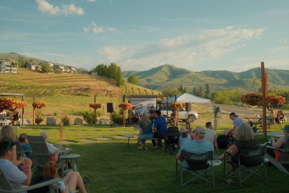 A small outdoor concert or performance taking place on a grassy lawn with an audience seated in folding chairs. Musicians perform under a white tent canopy with mountains and vineyard-covered hills in the background. Colorful hanging flower baskets line wooden posts, and residential buildings sit on the hillside. The setting is in a wine country or rural valley location during golden hour lighting.​​​​​​​​​​​​​​​​