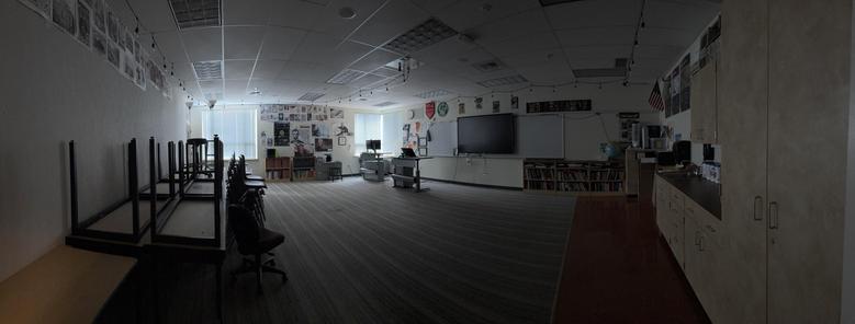 A panorama of an empty, dark classroom with all the desks stacked against one wall.